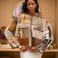 A woman modeling a patchwork quilted jacket with geometric and floral patterns in earthy tones of brown, beige, and white, paired with a white ribbed knit skirt in a warm boutique setting.