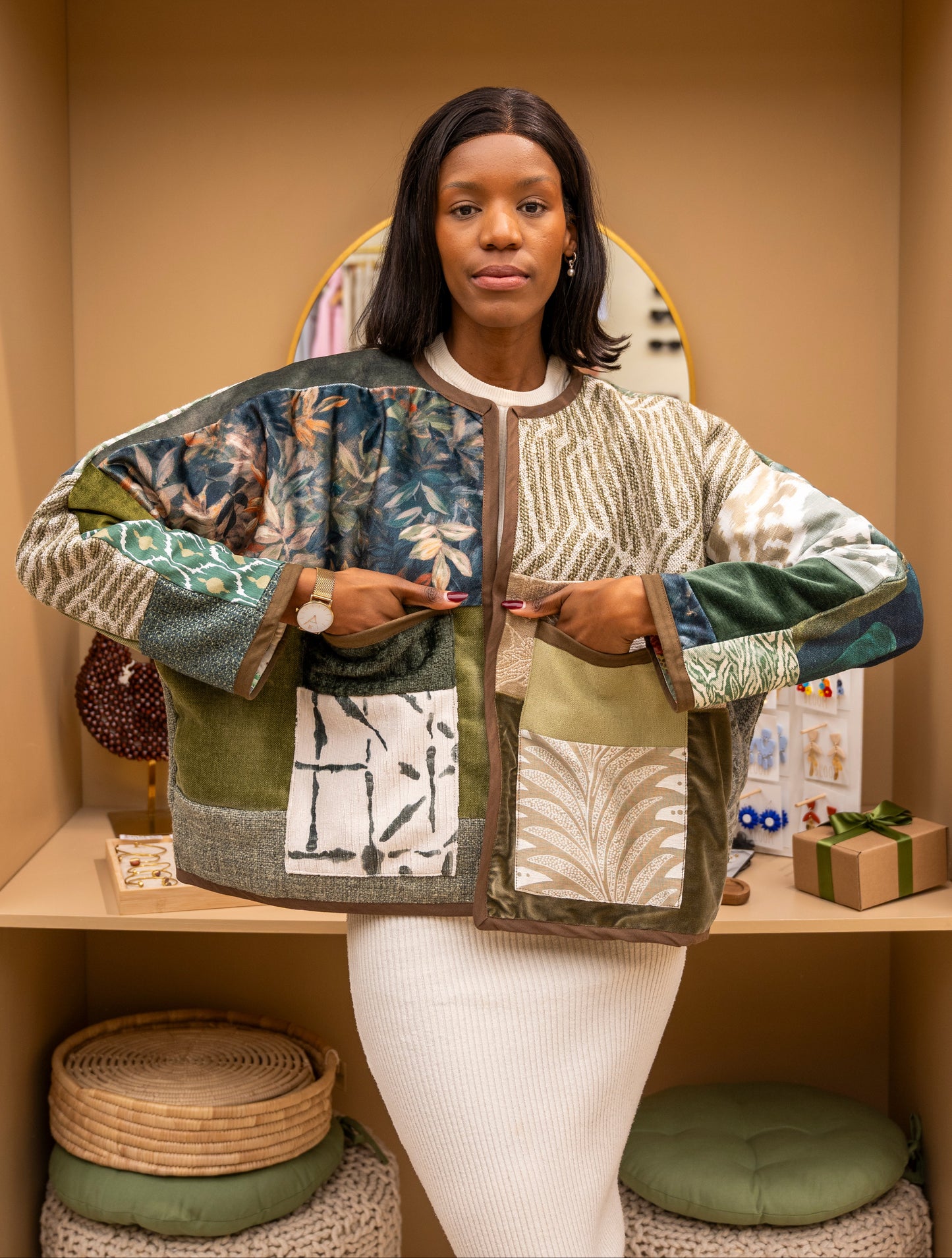 A woman stands in a warm indoor setting wearing a patchwork quilted jacket with mixed botanical and geometric patterns in beige, green, blue, and brown tones. The text 'Pause & Reflect' appears at the top of the image.