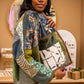A woman wearing a patchwork quilted jacket with floral and geometric patterns in green, blue, beige, and brown, posing in a boutique setting.