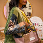 A woman in a boutique wearing a green velvet patchwork cardigan with patterned sleeves and a pink pocket, holding a matching pink and brown patchwork tote bag.